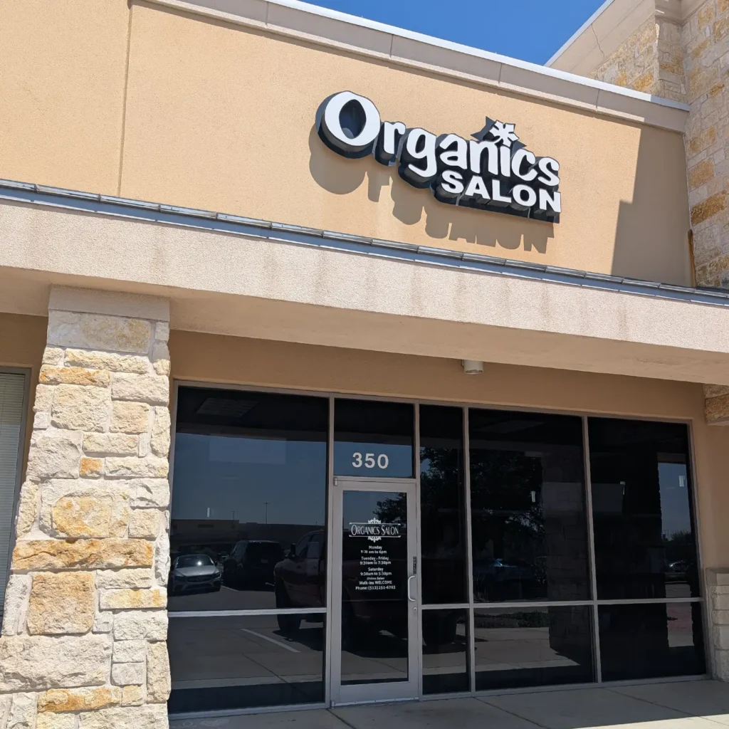 Storefront of Organics Salon with stone pillars, tan walls, and large windows. The salon’s name is displayed on a sign above the entrance. The door is marked with the number 350. - Pflugerville, TX