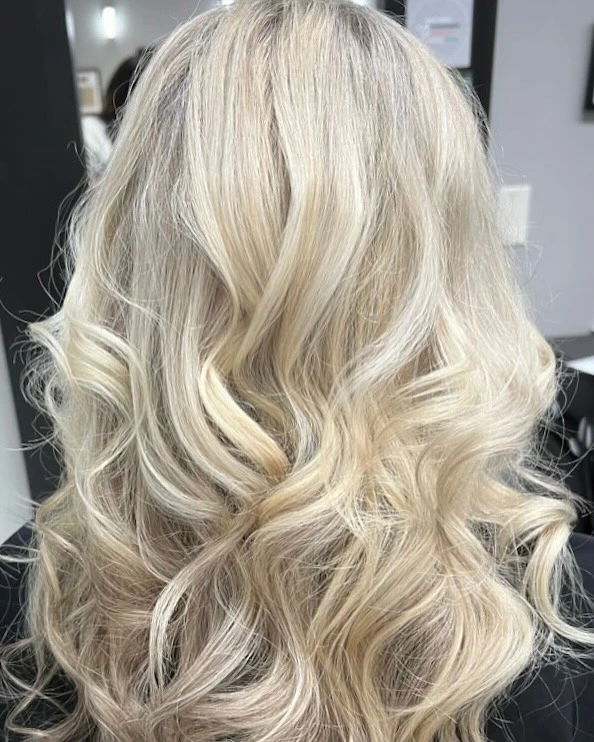 Back view of wavy, shoulder-length blonde hair with visible dark roots, photographed indoors in a salon setting. - Pflugerville, TX