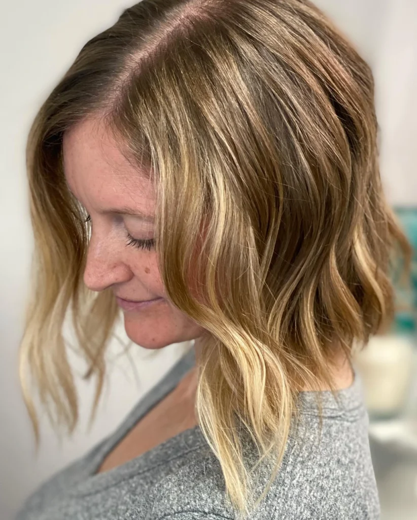 Person with shoulder-length, wavy, blonde balayage hair wearing a grey top, facing slightly left with eyes closed in a softly lit setting. - Pflugerville, TX