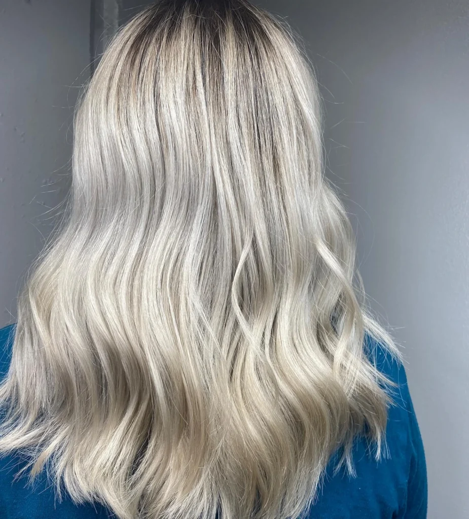 Person with shoulder-length, wavy platinum blonde hair facing away from the camera, wearing a teal top, against a plain light-colored background. - Pflugerville, TX