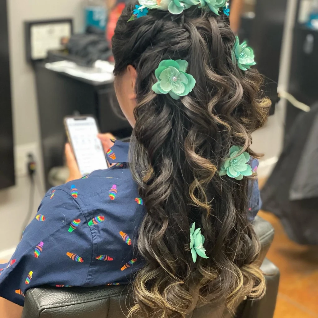Person with long, curly, dark hair adorned with green flower clips is sitting and looking at a smartphone, wearing a blue shirt with colorful print, in a salon setting. - Pflugerville, TX