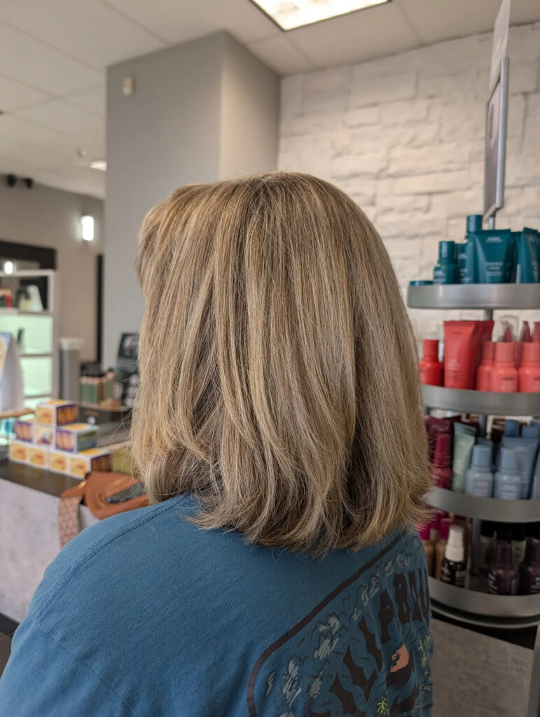 A person with shoulder-length, light blonde hair faces away from the camera inside a salon, with hair products visible on shelves in the background. - Pflugerville, TX