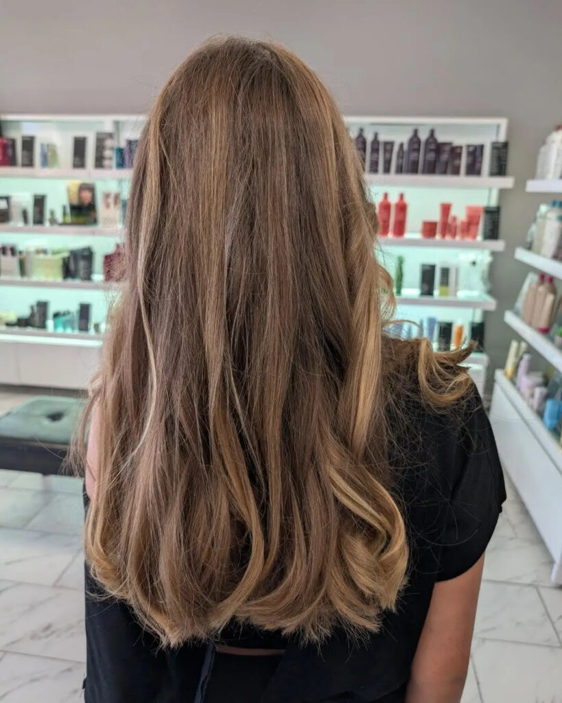 A person with long, light brown hair stands facing away in a salon, with shelves of hair products visible in the background. - Pflugerville, TX